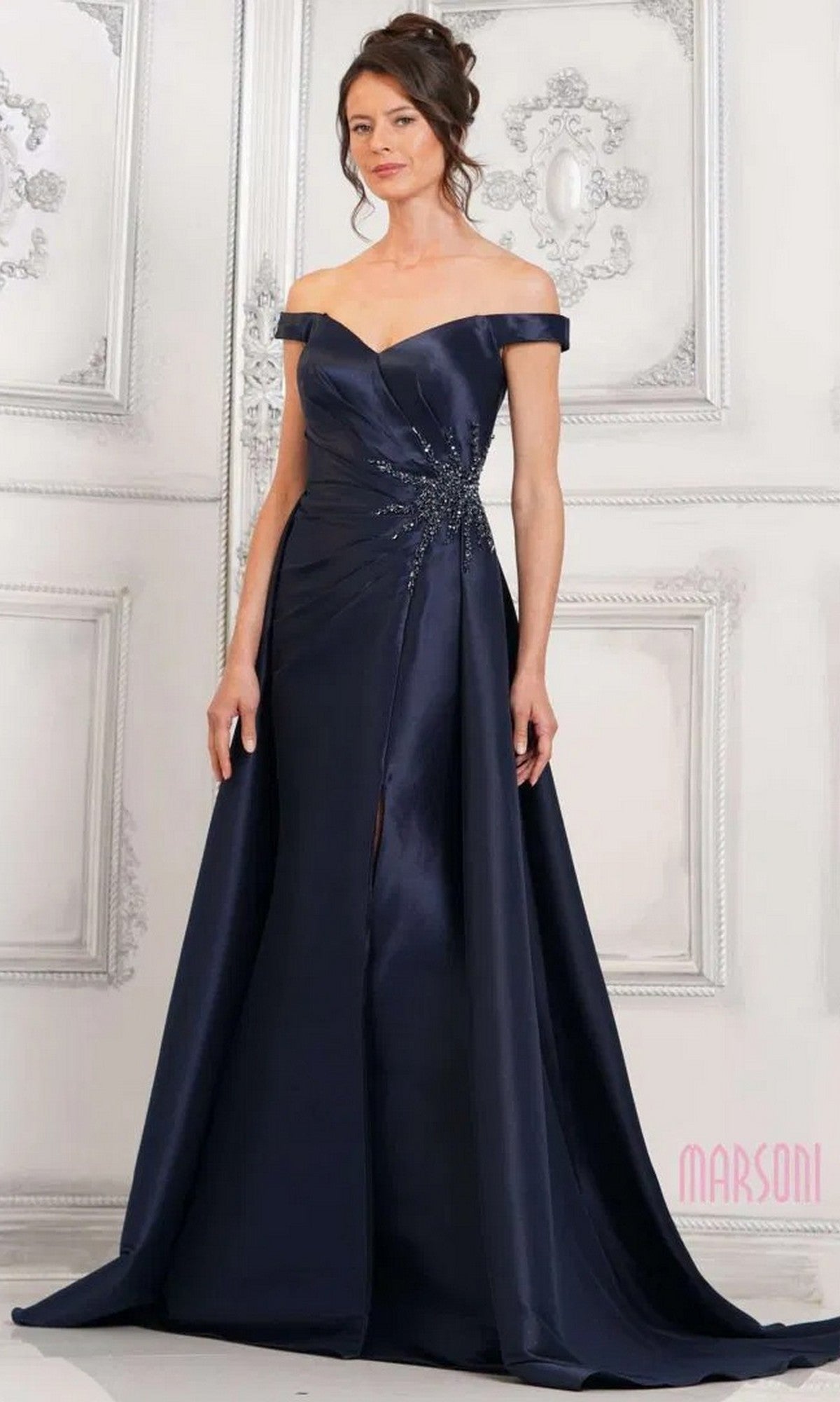 A woman wears the Marsoni MV1310 long formal dress, an off-the-shoulder navy blue gown with beaded embellishments and a mermaid silhouette, standing before an ornate white wall.
